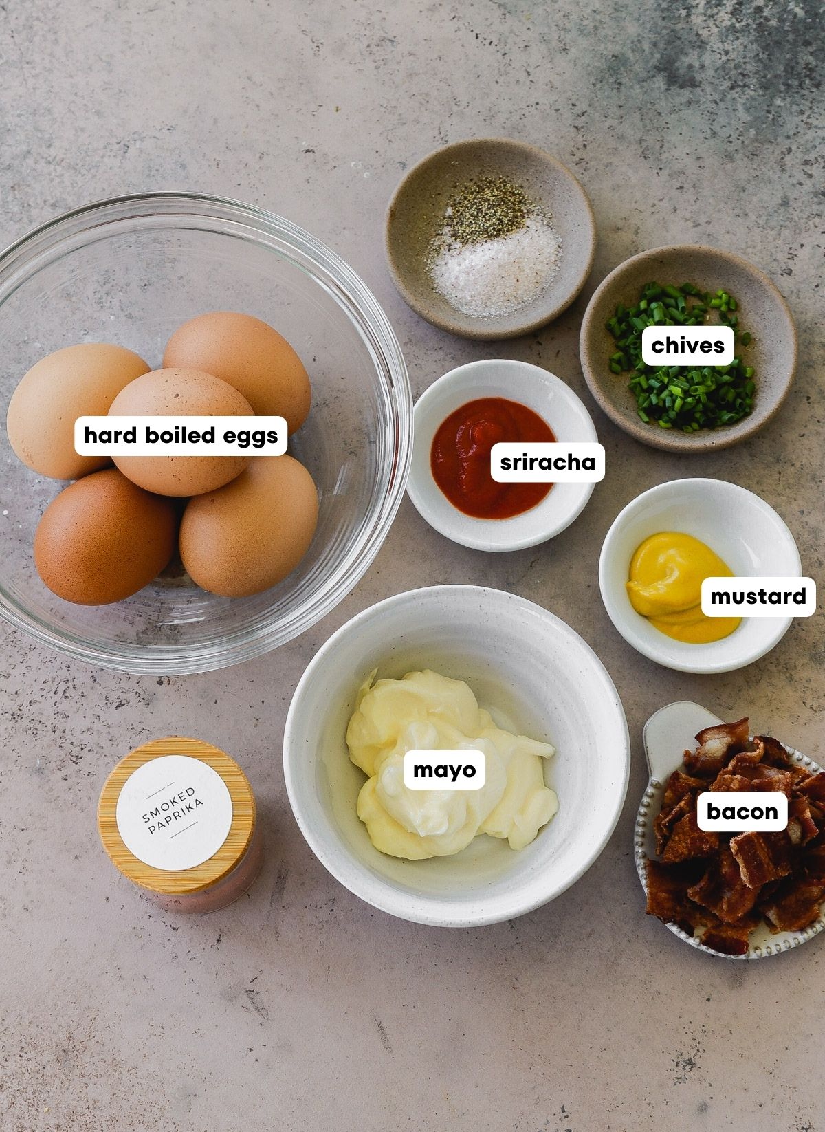 photo shot of all of the ingredients used to make sriracha deviled eggs with bacon: hard boiled eggs, mayo, sriracha, mustard, bacon, salt, pepper, paprika and chives.